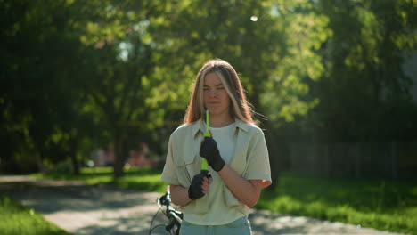 joven con cabello rubio se encuentra a la luz del sol, concentrándose mientras sostiene la bomba de aire verde cerca de la cara, sintiendo la brisa y el aleteo del cabello, el fondo muestra una escena tranquila del parque con árboles vibrantes