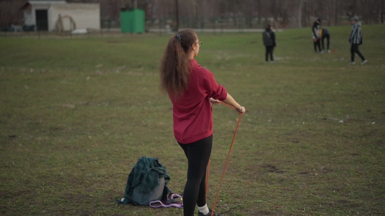 Woman Trains While Watching Game, Woman Performs Exercises In Park With Distant Players And Audience, Casually Dressed Woman Engages In Fitness Routines While Observing Activity In Park Setting
