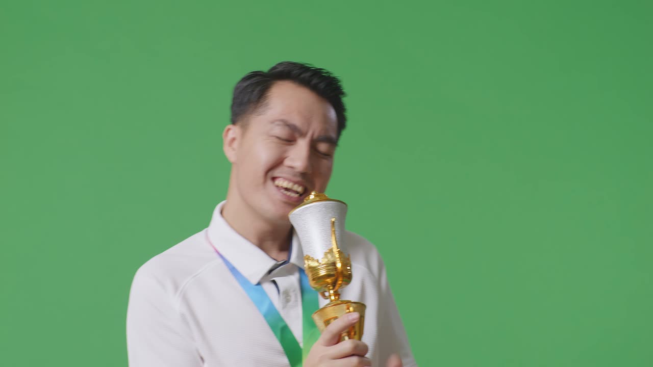 금메달을  아시아인 남자가 골드 트로피 위에서 노래를 부르며 우승을 축하하는 초록색 스크린 배경의 첫 번째 우승자입니다.