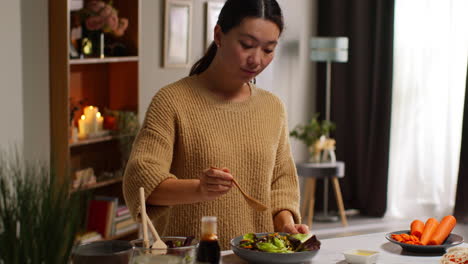 woman at home in kitchen preparing healthy vegetarian or vegan meal mixing in spoon of seeds into bowl of salad leaves