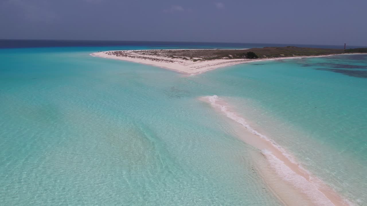 Ripresa aerea con allontanamento sopra un'isola di sabbia con onde di acqua oceanica azzurra e cristallina.
