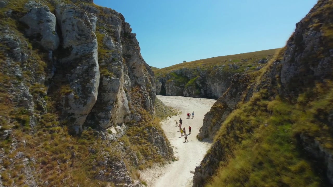 Drone narrow canyon rocky path hikers, cinematic passage. Low altitude aerial flight through deep limestone gorge with people trekking along the dry riverbed.