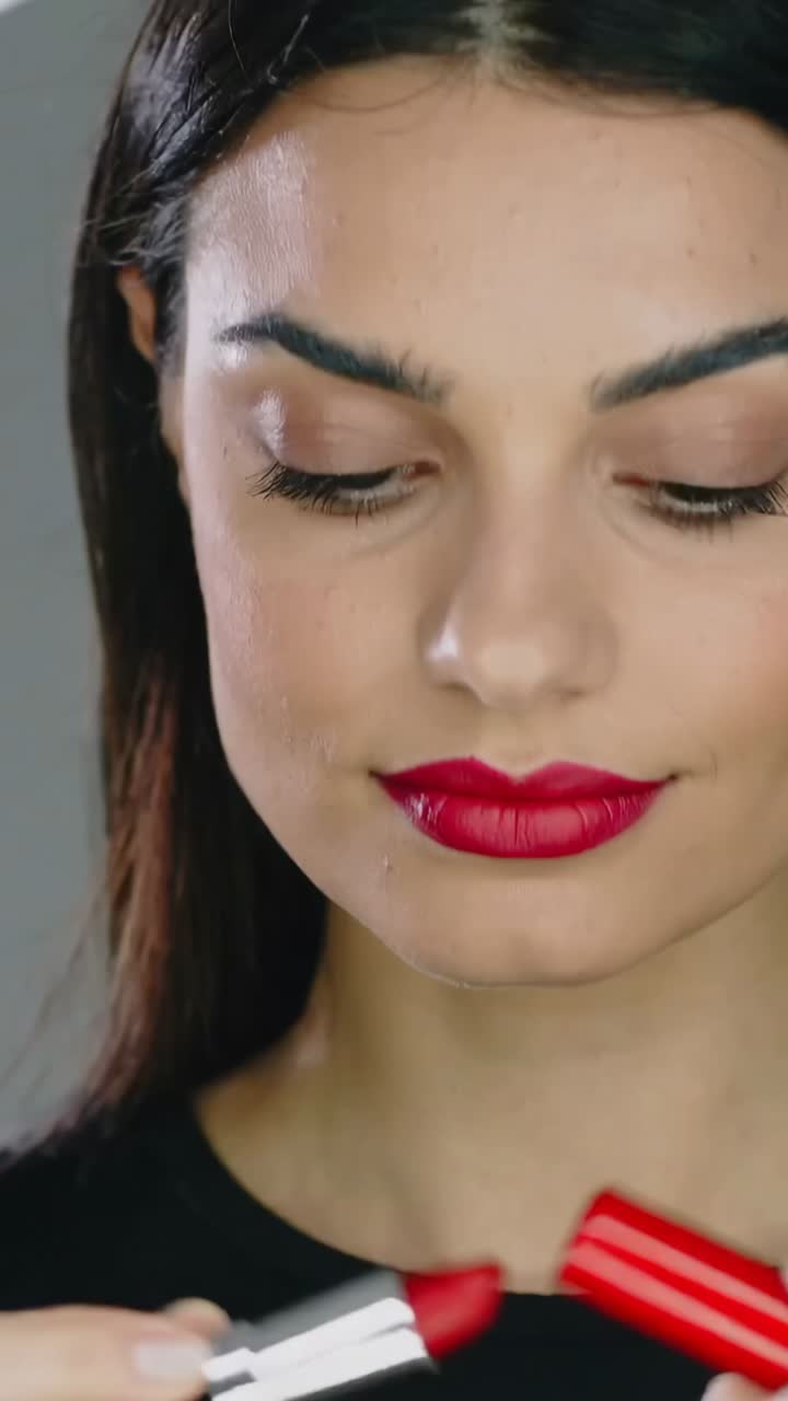 Vertical video: Bringing silver tube to lip, woman applying red lipstick in studio, creating gloss