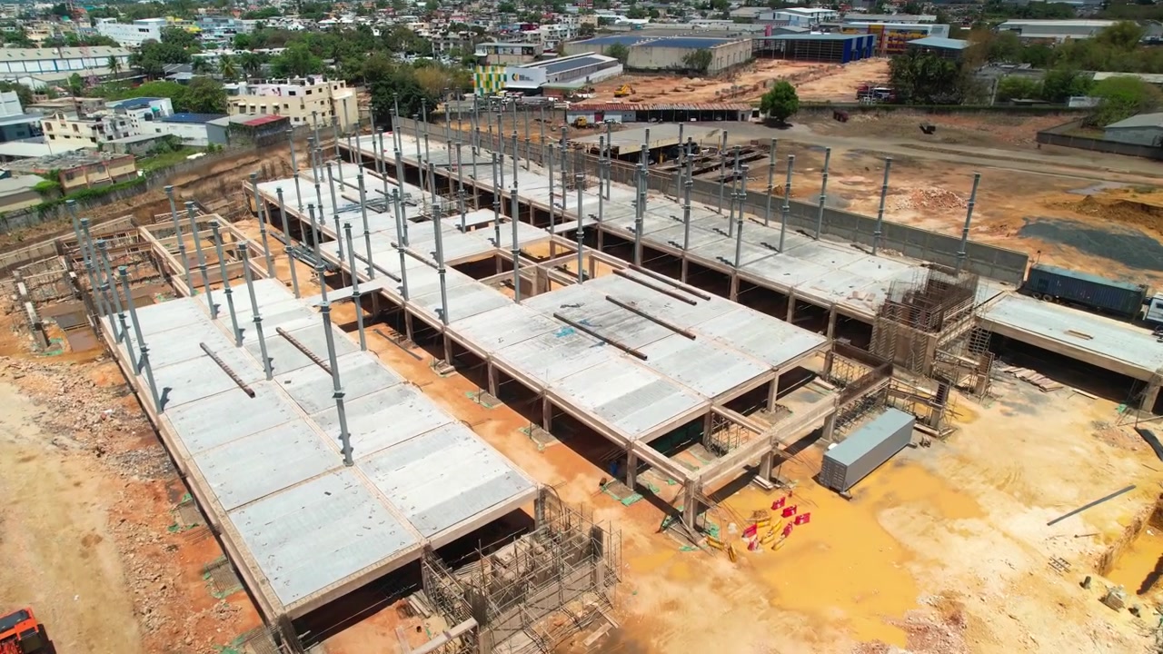 Imagem aérea de drone em ângulo alto, voando para frente e inclinando para baixo sobre um grande canteiro de obras de um edifício comercial, mostrando pilares de aço, lajes de concreto e arredores