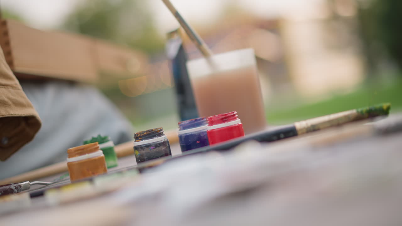 outdoor street art supplies closeup showing paint tubes, brushes, jar of water and palette arranged on pavement shallow focus highlights vibrant pigment and texture while blurred artist on bench