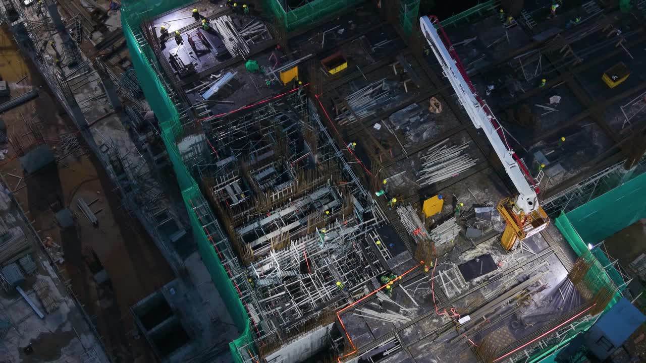 Slow aerial view of the VinHomes high-rise construction site near Hanoi's West Lake at night. Bright work lights illuminate workers and concrete pumping activity. Development and labor