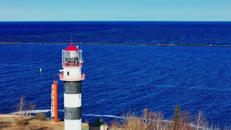 a tall black and white lighthouse stands near bare trees facing bright blue baltic sea and sandy shore stretching into the distance under a clear sky.