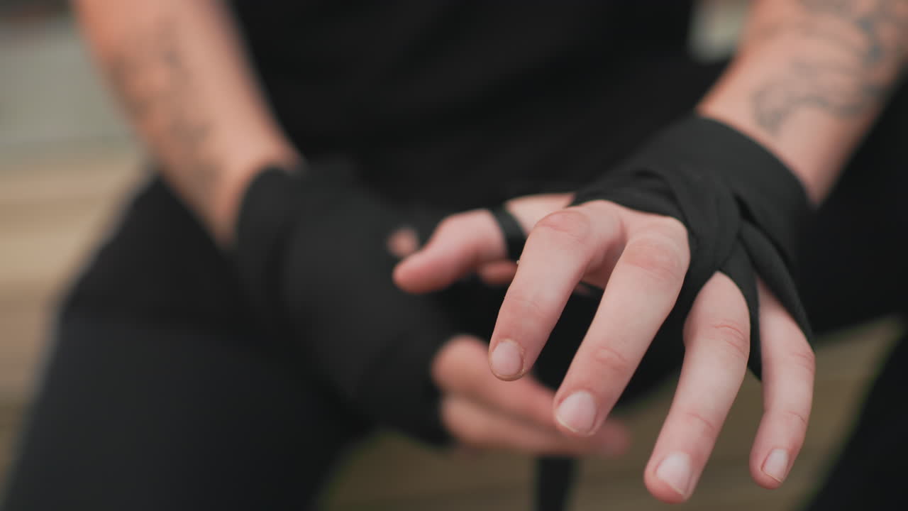 Un combattente caucasico di MMA che si assicura i bendaggi neri sulle mani seduto su una panchina, primo piano degli avambracci tatuati e delle dita precise che infilano la cinghia, calma routine tattica prima dello sparring o dell'incontro, tecnica mirata