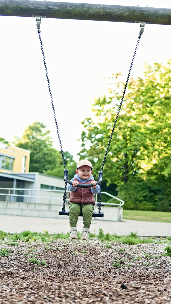 Toddler in blazer sits on swing in city park. Boy swings on swing on playground. Child plays and has fun on street. Children's swing