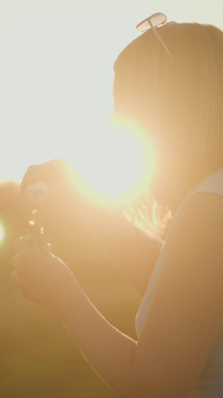 머리에 선글라스를 착용한 파란색 수갑없는 상자를 입은 여성은 황금 시간 동안 거품을 불어넣고, 빛이 그녀의 머리카락을 부드럽게 비추고, 부드러운 흐릿한 자연 배경과 함께 아름다운 가장자리 빛을 만니다.