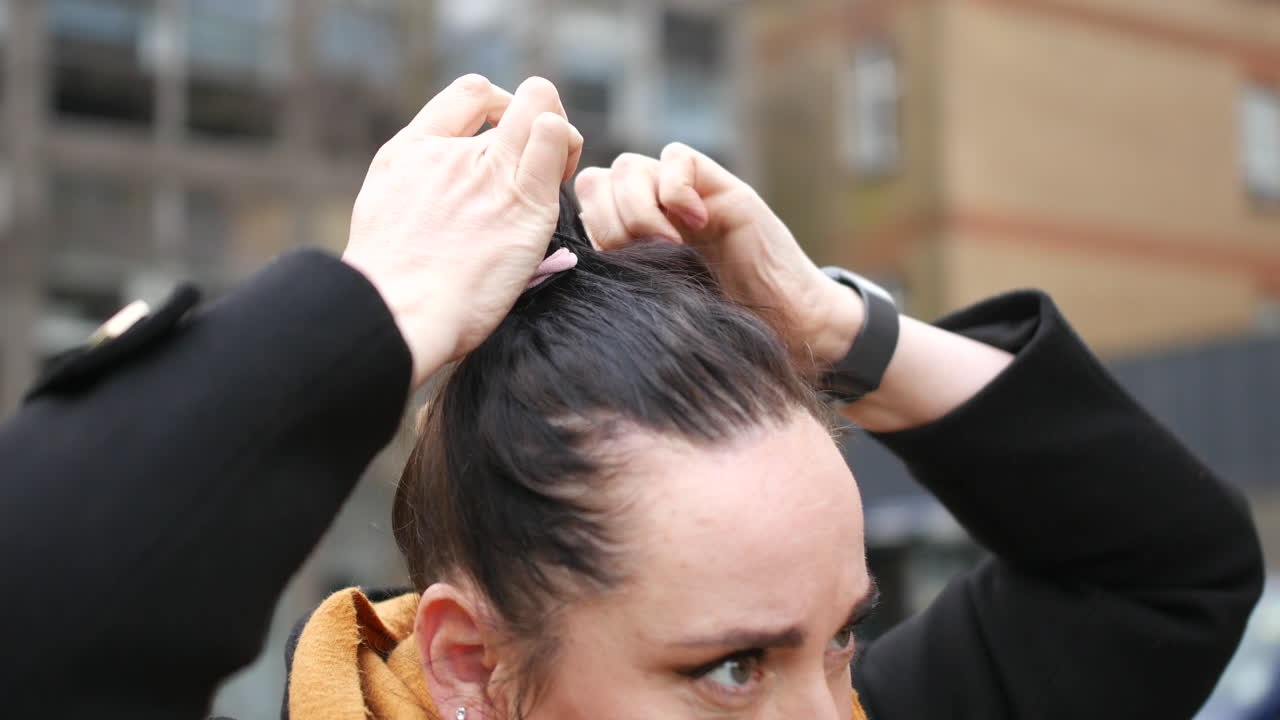 Mulher prendendo o cabelo escuro em um rabo de cavalo com um elástico enquanto está ao ar livre em ambiente urbano. Close-up capturando as mãos ajustando o penteado, vestindo casaco preto e cachecol mostarda perto de edifícios modernos.