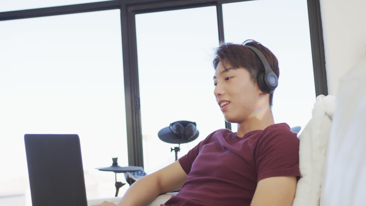 행복한 아시아 남성 십대가 헤드폰을 착용하고 거실에서 노트북을 사용합니다.