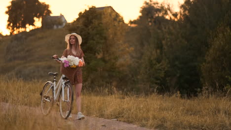 Young-beautiful-blonde-girl-in-summer-in-dress-and-hat-walking-on-road-with-bike-and-flowers-in-slow-motion.