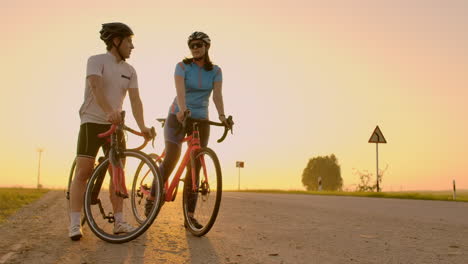 A-man-and-a-woman-in-helmets-with-bicycles-stand-and-talk-at-sunset.-Rest-after-a-bike-ride-on-the-highway.-Track-bikes.-Couple-sports