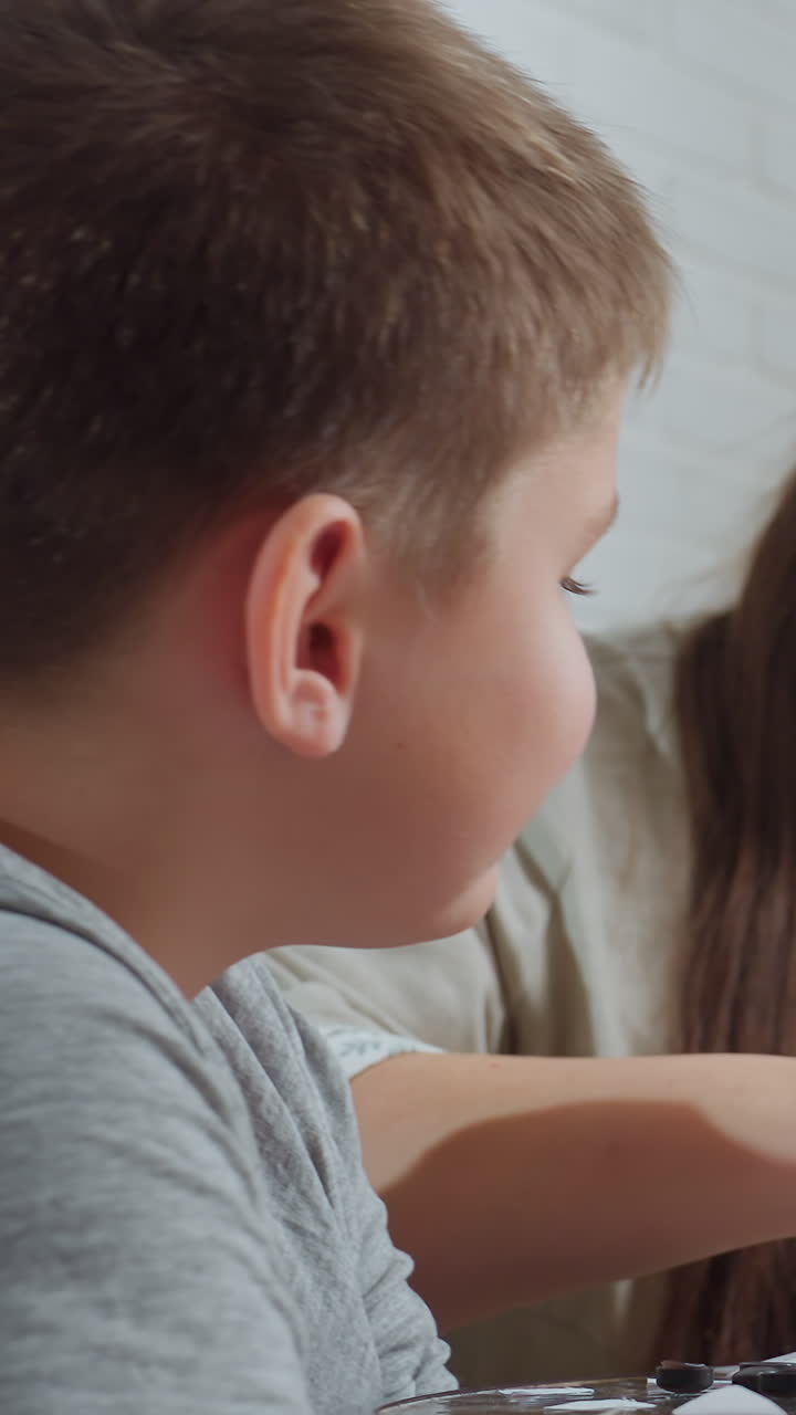 탁자에서 장식품을 만드는 어린 남자아이를 지도하는 백인 엄마, 직접적인 지도와 부드러운 격려, 배경에는 크리스마스 트리 조명, 학습과 놀이의 친밀한 공유 순간