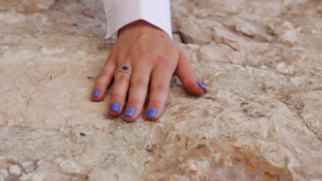 Young-girl-wearing-a-white-long-sleeve-shirt-with-a-blue-ring-and-blue-nails-runs-her-hand-gently-along-a-textured-rocky-wall