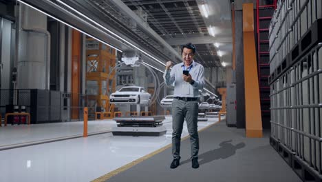 cuerpo lleno de un trabajador profesional masculino asiático de pie con su teléfono inteligente en la fábrica de automóviles ev, instalación industrial de alta tecnología, levanta el puño con el objetivo de gritar