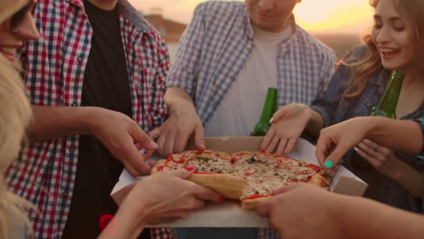 a company of six young people parses pieces of hot pizza and eats together. this is a rooftop party with a beer.