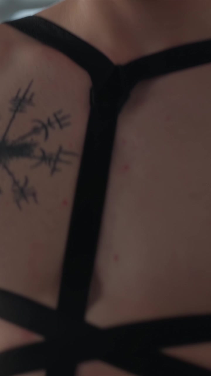 Close-up de homem com tatuagens e óculos de sol, usando algo com alças. Mãos manipulam algemas perto da cintura de outro homem. O fundo claro sugere um espaço interior moderno com grandes janelas.