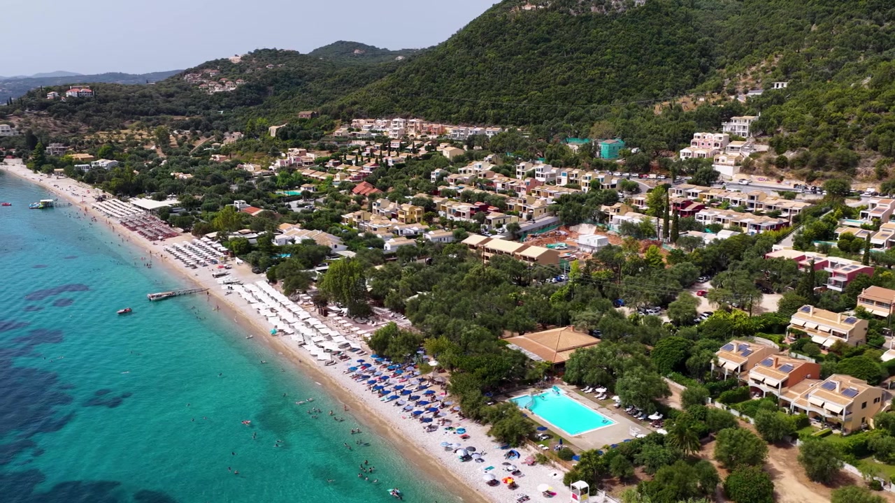 Ampla imagem aérea da praia de Barbati em Corfu, Grécia, água do mar turquesa cristalina, montanhas verdes exuberantes, espreguiçadeiras ao longo da paisagem costeira Jônica