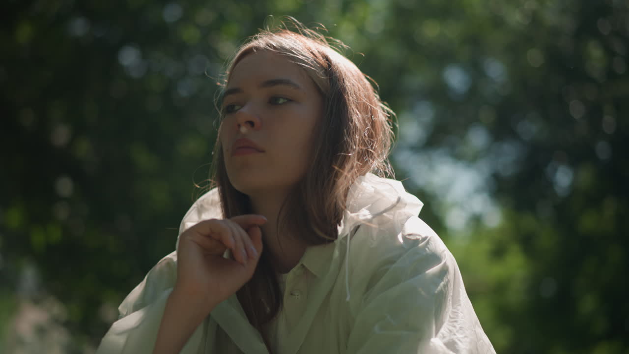 joven con impermeable translúcido baja la capucha y descansa la cabeza sobre la mano pensativamente, con la luz del sol reflejándose suavemente en su cara y la vegetación exuberante en el fondo