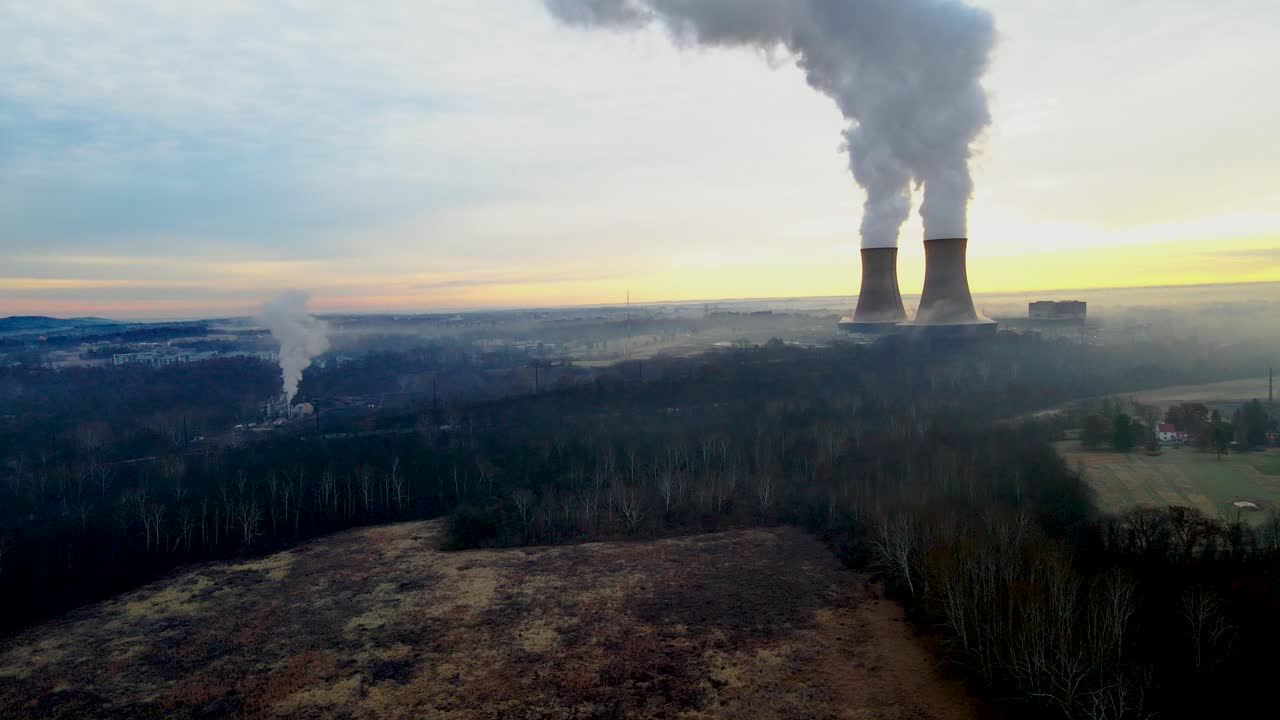 Un impianto nucleare all'alba, con le torri di raffreddamento che sbuffano vapore nel cielo mattutino. Movimento lento della telecamera in dolly out.