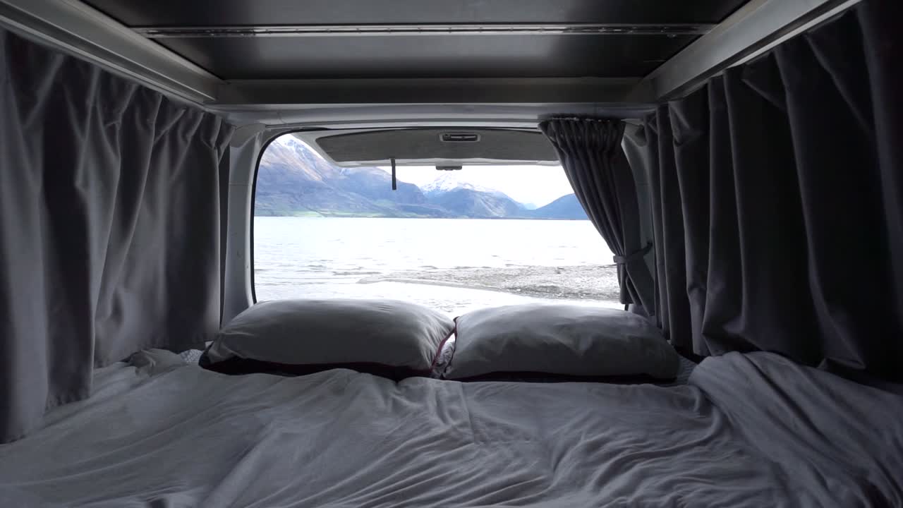 cámara lenta - interior de la cama de la casa rodante con almohadas y mantas junto al lago wakatipu, queenstown, nueva zelanda con montañas cubiertas de nieve en el fondo