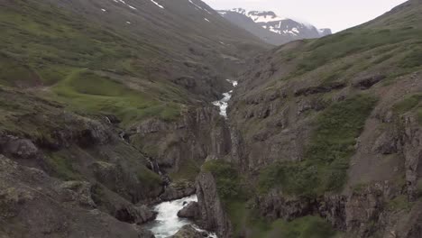 Rugged-rocky-cliffs-surround-whitewater-river-in-Fagridalur-valley,-aerial