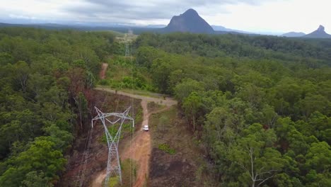 aerial drone shot away from a white car, bypassing power lines, on sand, dirt road, surrounded by forest, at glass house mountains national park, in queensland, australia