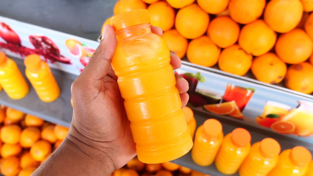 una mano sosteniendo una botella de jugo de naranja frente a un estante lleno de naranjas