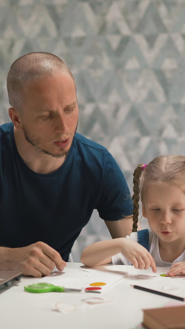 shaved bald father helps glue colorful sticker on paper sheet to pretty little daughter sitting at white table with laptop in room at isolation