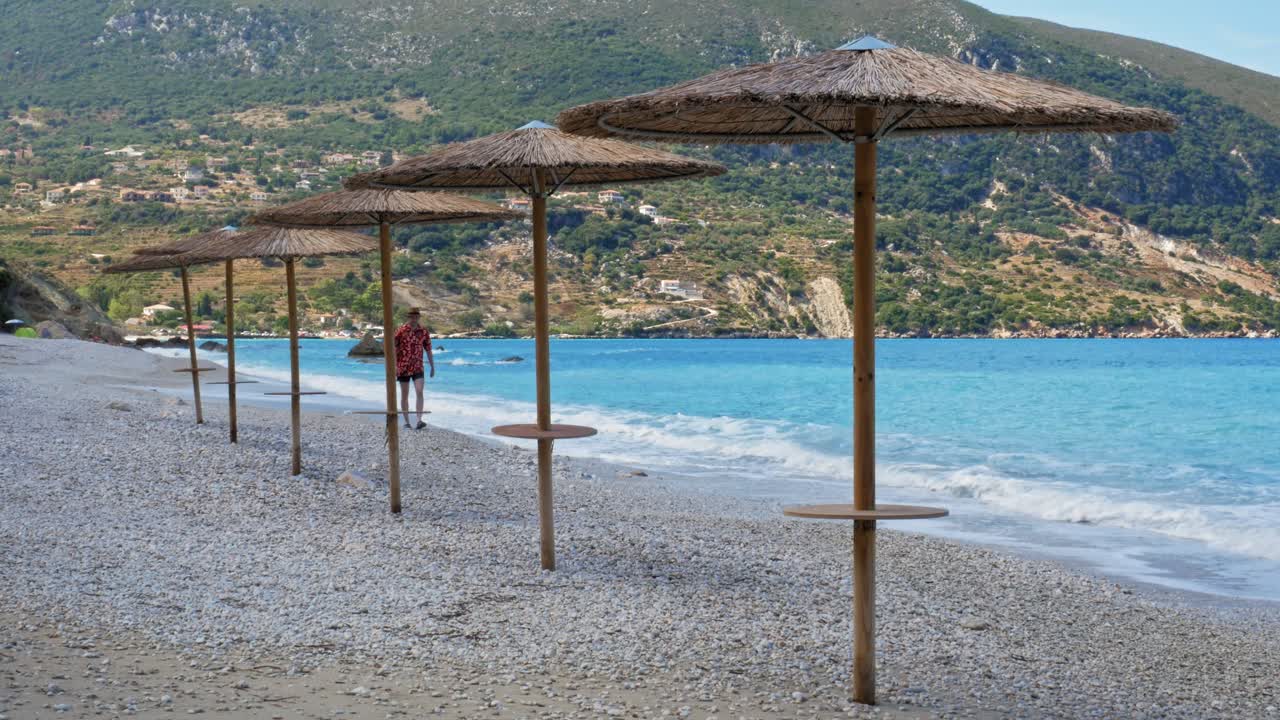 A Male Tourist Relaxing Walking By The Beautiful Shore With Native Umbrellas And Mountains Background At Agia Kiriaki Beach Near Zola Village, Kefalonia, Greece
