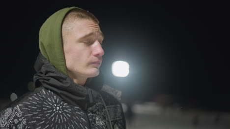 side view of student dressed in hooded winter jacket walking outdoors on cold night with visible vapor escaping mouth, soft glow from streetlights and blurred background setting wintry mood