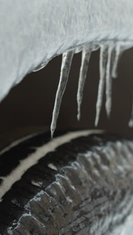 close-up of car tire covered with snow and hanging icicles during winter, featuring a blurred background with the rear view of another car