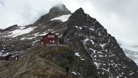 a young, fit man hiker with long hair is standing on the edge of the cliff next to dossen hut on top of the big mountain with the peak and clouds behind him