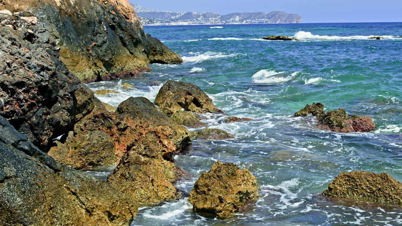 Onde che si infrangono contro una costa rocciosa mentre barche navigano in lontananza sotto un cielo azzurro brillante vicino a una comunità costiera.