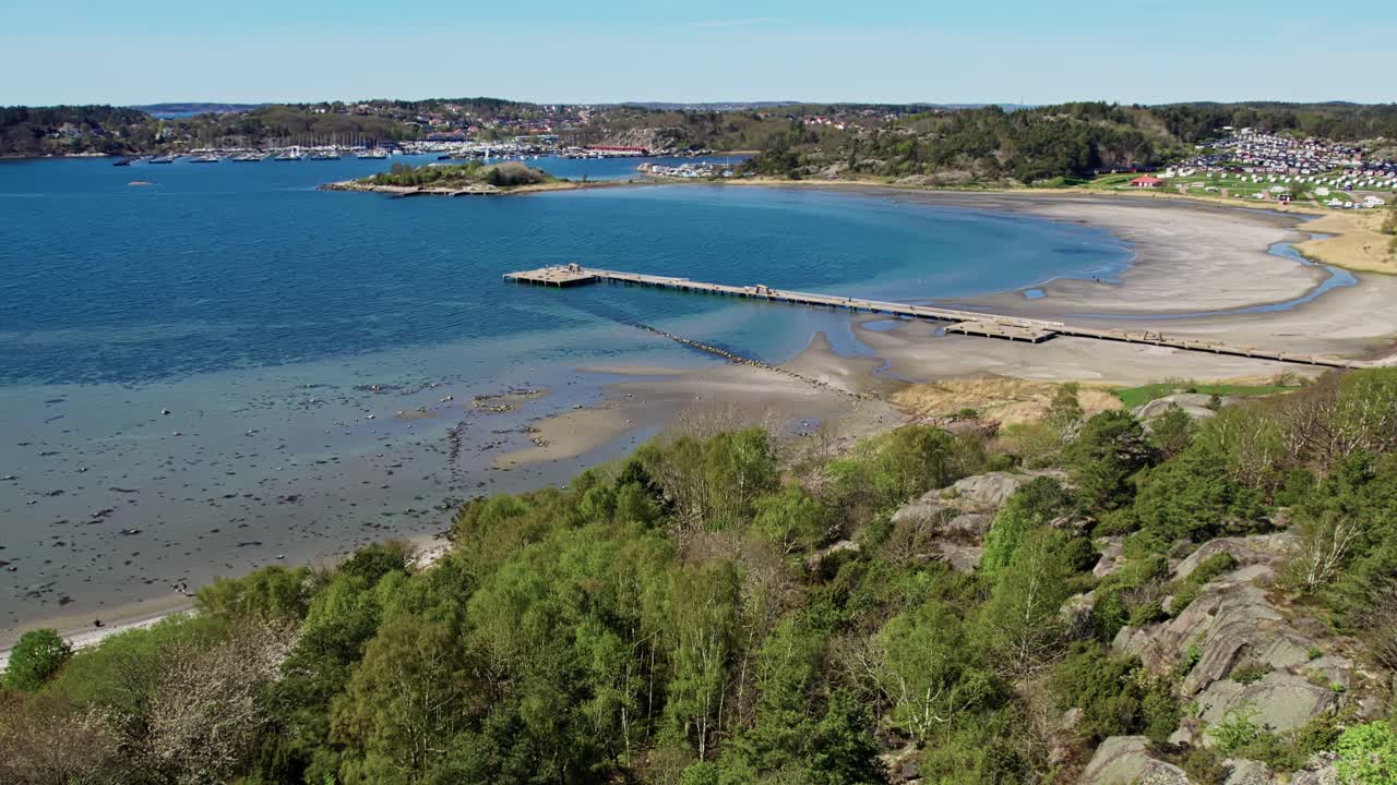 Slow panning aerial footage of Askimsbadet, a sandy beach south of Gothenburg, Sweden. Highlights the wide shoreline, shallow tidal waters, and the 259 meter long pier into the blue sea