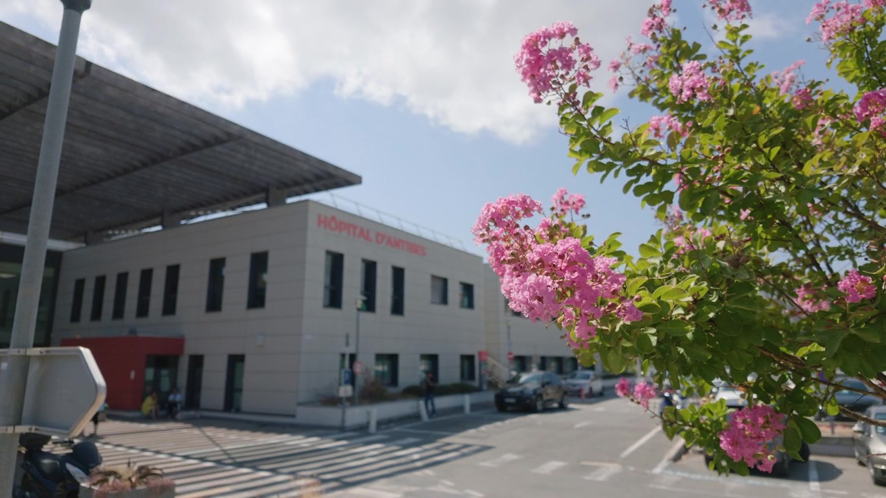 Edifício do Hospital de Antibes com uma faixa de pedestres, carros estacionados e flores de resedá rosa sob um céu azul nublado, Antibes, França