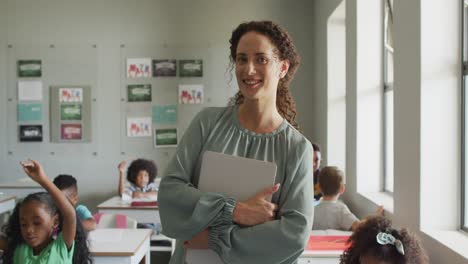 video of happy caucasian female teacher holding laptop in class of diverse pupils