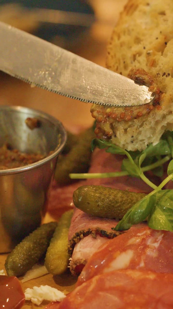 A knife spreads grain mustard onto a slice of rustic bread above a charcuterie board with cured meats, pickles, and microgreens in warm, ambient lighting