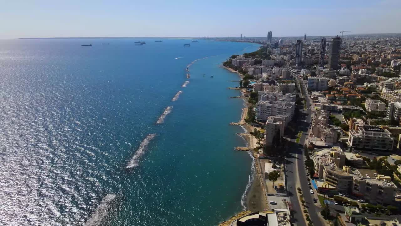 Drone video shows the sweeping coastline of Durrës, Albania, with blue waters, urban sprawl, and protective breakwaters under a clear sky, capturing vibrant city life and serene sea