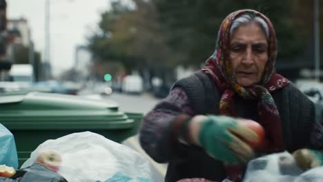 an elderly woman in a warm coat and headscarf sifts through trash bags on a busy city street. she carefully looks for recyclable items amid the discarded waste.