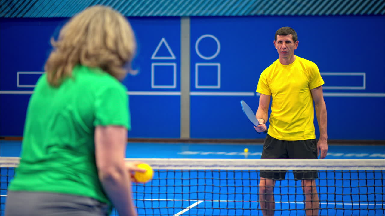 A man and a woman playing pickleball on a blue, inside court
