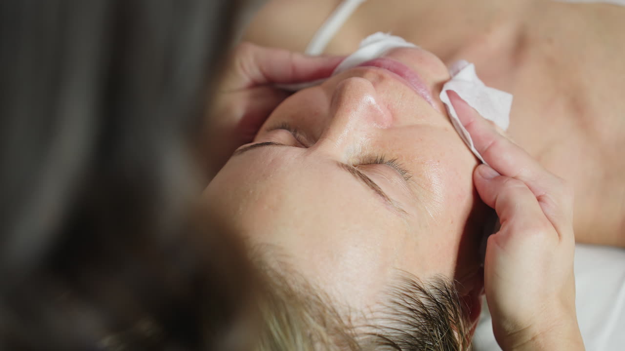 Individual on white sleeveless cloth lies with eyes closed while facialist uses wet wipes to gently cleanse face, removing product residue and impurities, highlighting hygienic skincare routine