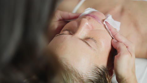 individual on white sleeveless cloth lies with eyes closed while facialist uses wet wipes to gently cleanse face, removing product residue and impurities, highlighting hygienic skincare routine