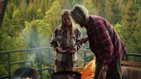 Ein-Blondes-Mädchen-Hilft-Ihrem-Blonden-Freund,-Würstchen-Vom-Grill-Auf-Dem-Balkon-Eines-Landhauses-Mit-Blick-Auf-Den-Wald-Und-Die-Berge-Zu-Nehmen