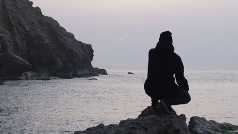 man sitting on rough rock watching sunrise as seagulls fly over sea, fascinated by beautiful view