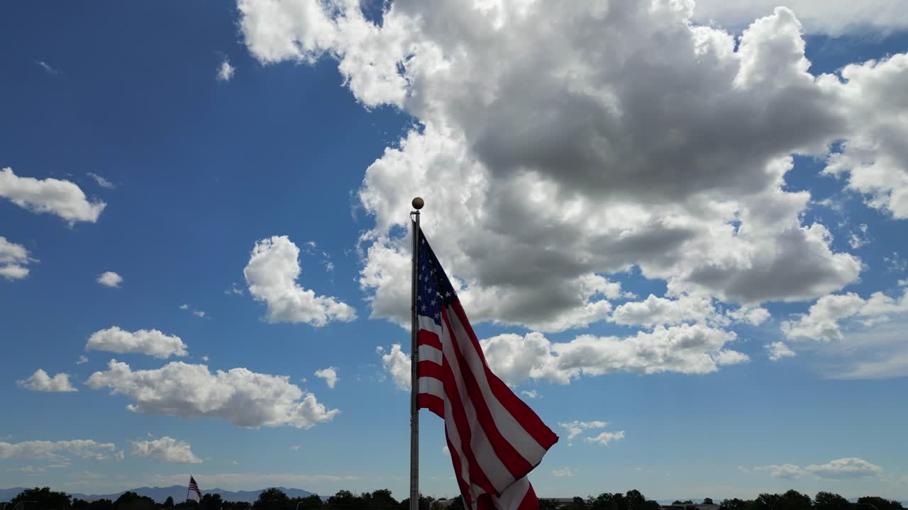 American flag USA blowing waving in the wind on beautiful sunny summer day with clouds and blue skies as drone flys around flagpole with small town cresting in bottom of shot - in 4K 60fps