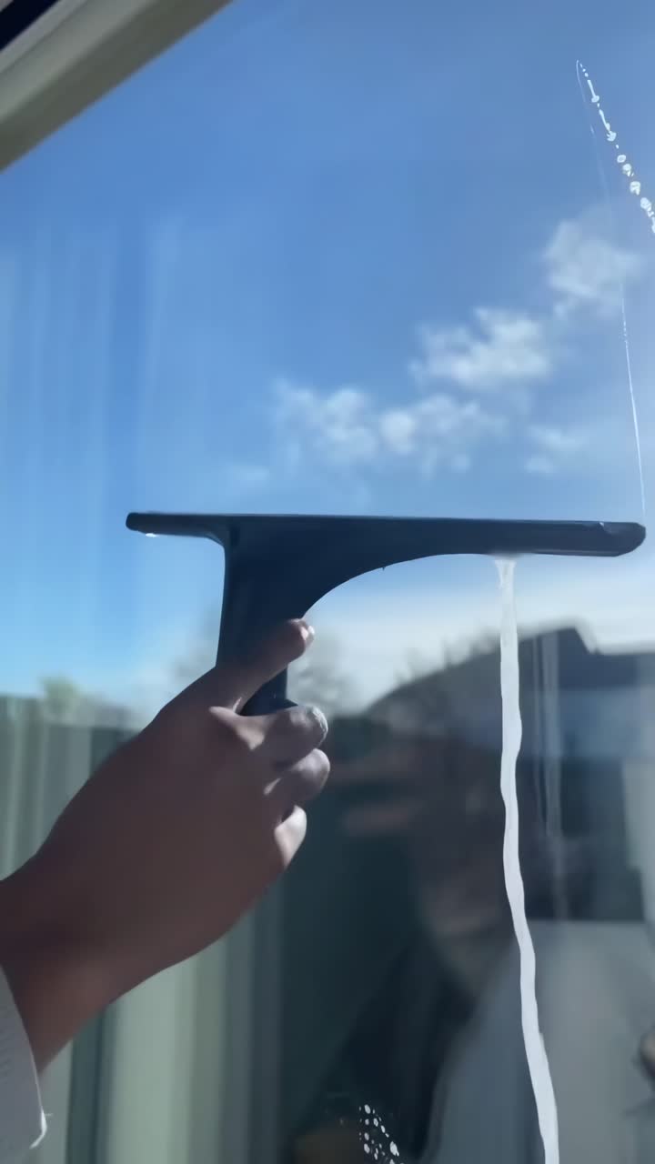 Video ravvicinato, con inquadratura dal basso di una mano che pulisce una finestra con un tergivetro contro un cielo soleggiato.