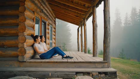 two friends are sitting on the wooden porch of a cozy log cabin, taking in the serene atmosphere of a misty mountain morning, enjoying the peace and quiet of nature